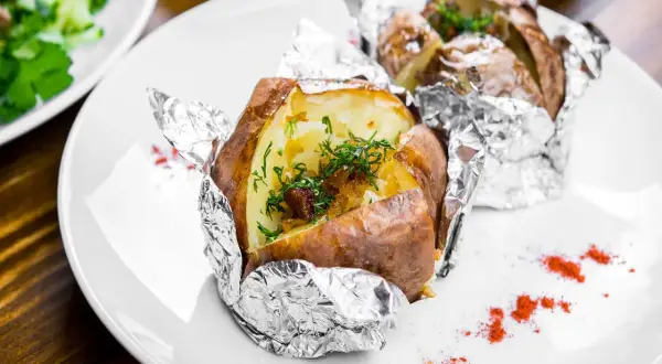 Patate al cartoccio fumanti, condite con erbe fresche e cipolla caramellata, servite su un piatto bianco con un tocco di paprika.