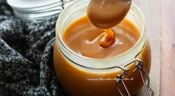 Un cucchiaio raccoglie un filo di caramello salato perfetto, denso e dorato, da un barattolo di vetro.
