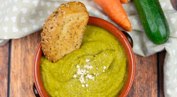 Una ciotola di cremosa crema di zucchine, guarnita con fiocchi d'avena e accompagnata da pane integrale tostato. Carote e zucchine fresche sullo sfondo.