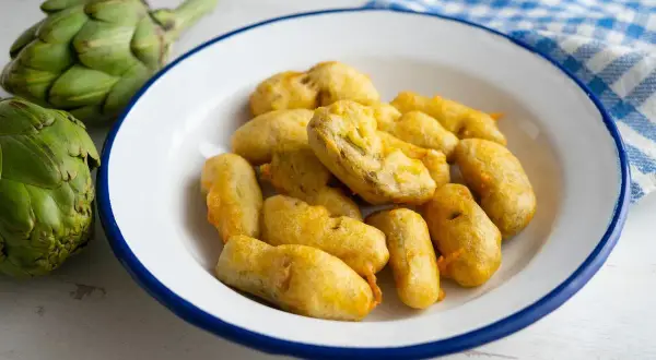 Carciofi fritti alla romana, dorati e croccanti, preparati con una pastella leggera. Accanto, carciofi freschi e un tovagliolo a quadretti.