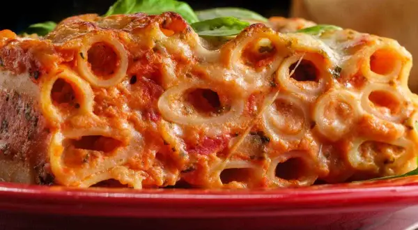Un primo piatto invitante di ziti al forno, con sugo rosso e formaggio filante, guarnito con foglie di basilico fresco.