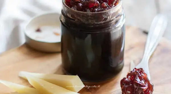 Un vasetto di marmellata di cipolle rosse, con un cucchiaino che ne assaggia un po' su una fetta di formaggio.