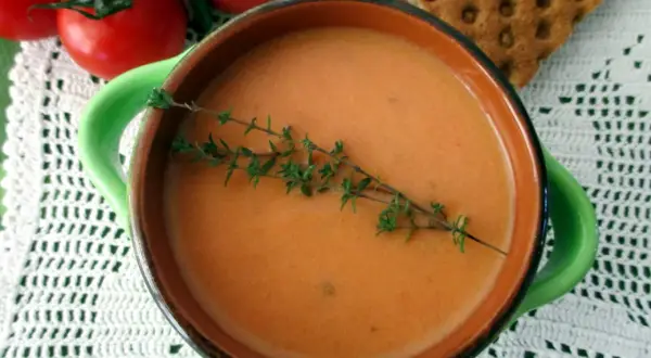 Una ciotola di cremosa crema di pomodoro, guarnita con timo fresco, accanto a pomodori maturi e cracker croccanti.