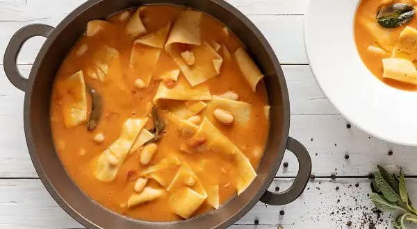 Un saporito piatto di maltagliati con fagioli, servito in una padella scura e una ciotola bianca, con erbe aromatiche e pepe nero a guarnire.