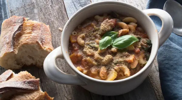 Una ciotola di pasta e fagioli in scatola, guarnita con basilico fresco e parmigiano, accanto a pane rustico su un tavolo di legno.