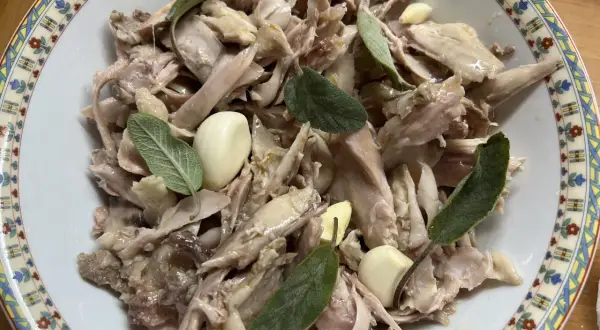 Piatto di tonno di coniglio sfilacciato, condito con aglio e foglie di salvia fresca, servito in una ciotola decorata.