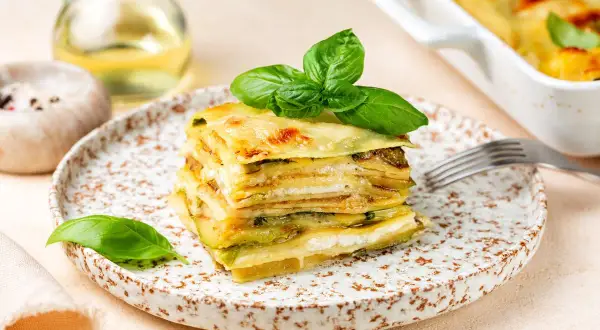 Una fetta di parmigiana di zucchine bianca alla napoletana, stratificata e dorata, guarnita con basilico fresco.