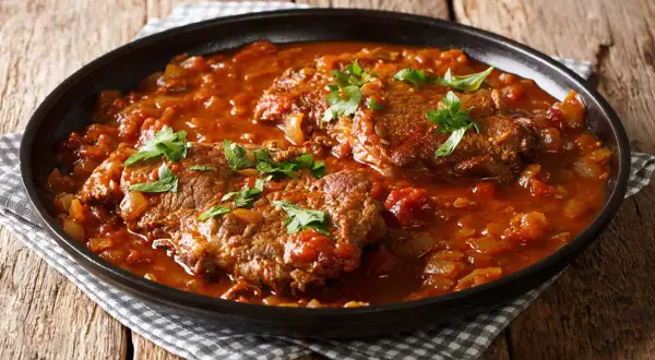 Carne alla pizzaiola morbida, succulenta e profumata, servita in un piatto scuro su un tavolo di legno.