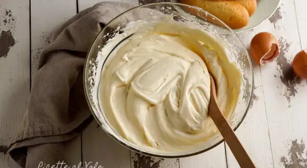 Una ciotola di soffice crema al mascarpone, pronta per preparare un delizioso tiramisù. Accanto, savoiardi e gusci d'uovo.