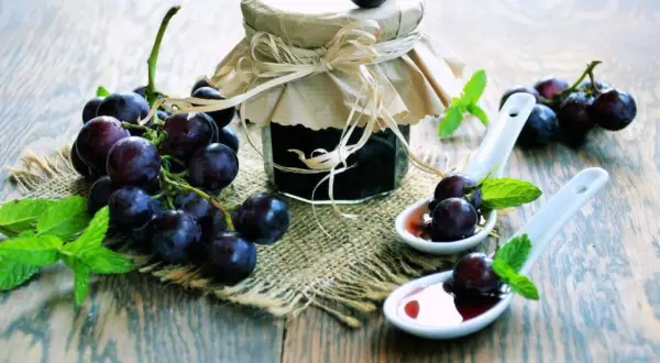 Marmellata di uva con buccia, con grappoli d'uva fresca e foglie di menta su un tavolo di legno.