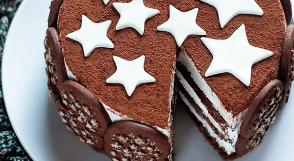 Torta Pan di Stelle ricoperta di cacao, decorata con stelle bianche e biscotti al cioccolato. Una fetta mostra gli strati soffici.