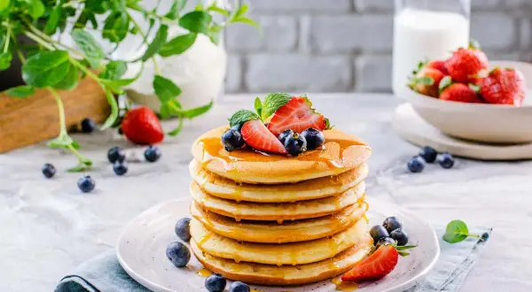 Pila di soffici pancake senza glutine e lattosio, guarniti con fragole, mirtilli e sciroppo d'acero.