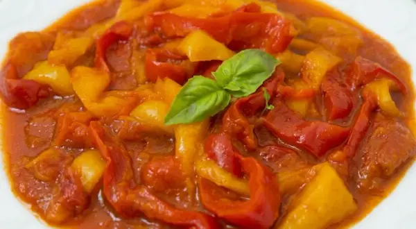 Peperonata ricetta della nonna: peperoni rossi e gialli cotti lentamente in un sugo saporito, guarniti con una fogliolina di basilico fresco.