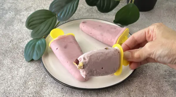 Tre ghiaccioli rosa, un gelato allo yogurt fatto in casa, su un piatto bianco con foglie verdi sullo sfondo.