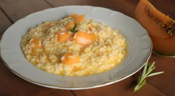 Un cremoso risotto con pezzi di melone e un rametto di rosmarino, accanto a una fetta di melone.