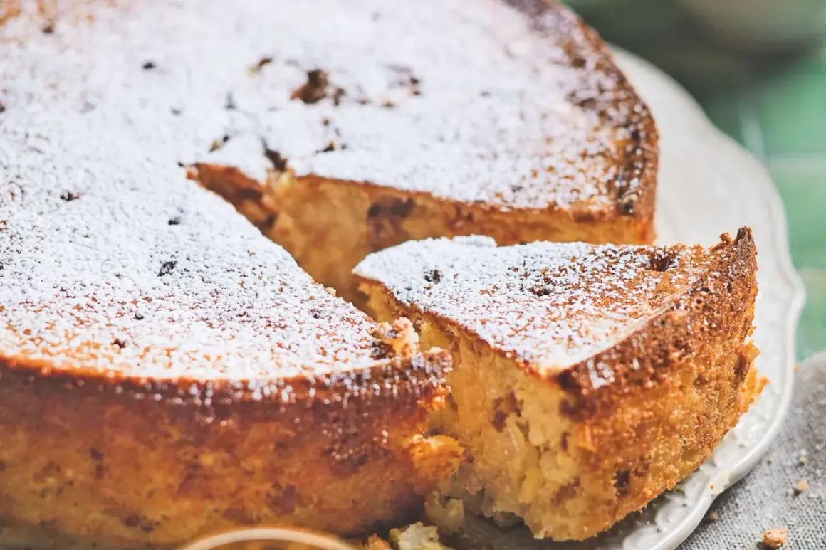 Torta di riso ricetta originale, soffice e dorata, cosparsa di zucchero a velo. Una fetta mostra la consistenza cremosa.