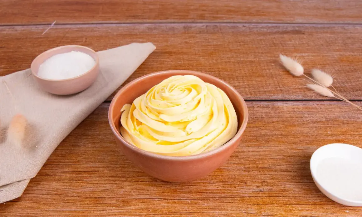 Una ciotola piena di soffice crema al burro, pronta per decorare torte. Accanto, ciotoline con zucchero e decorazioni naturali.