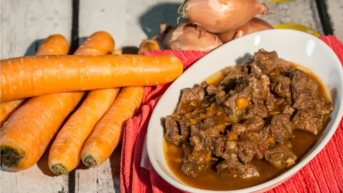 Un invitante spezzatino di manzo in un piatto bianco, con carote fresche e cipolle sullo sfondo.