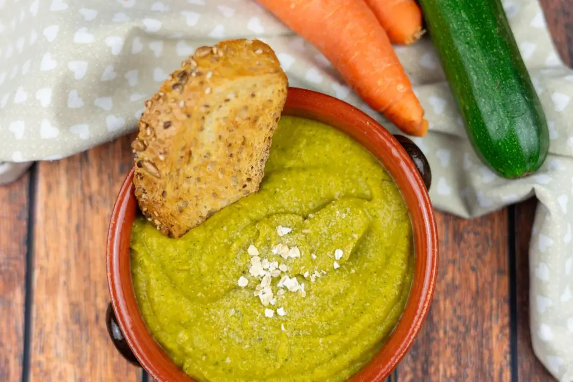 Una ciotola di cremosa crema di zucchine, guarnita con fiocchi d'avena e accompagnata da pane integrale tostato. Carote e zucchine fresche sullo sfondo.