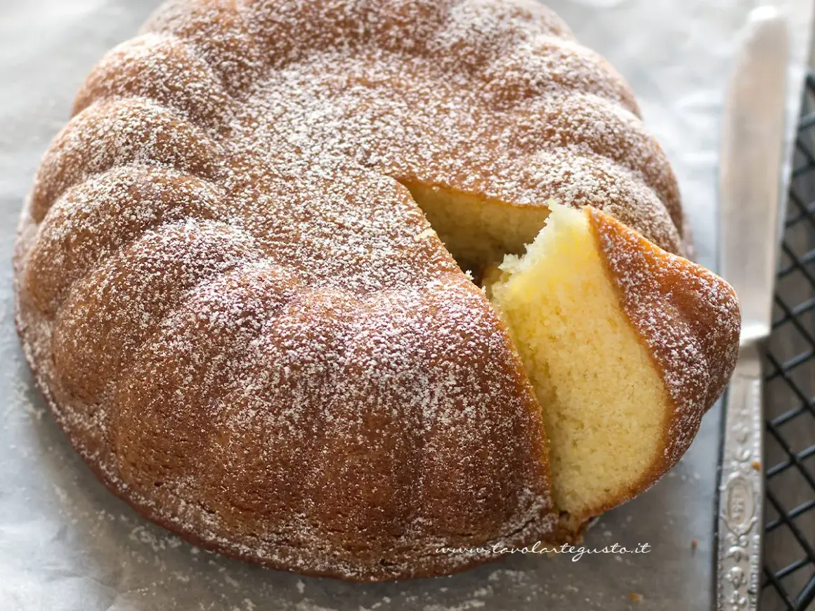 Torta 7 vasetti soffice e dorata, cosparsa di zucchero a velo. Una fetta è stata tagliata, rivelando l'interno profumato.