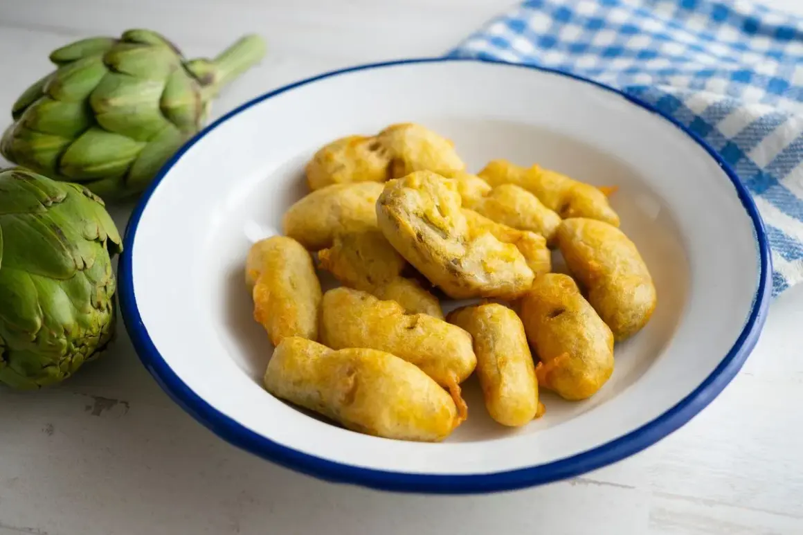 Carciofi fritti alla romana, dorati e croccanti, preparati con una pastella leggera. Accanto, carciofi freschi e un tovagliolo a quadretti.