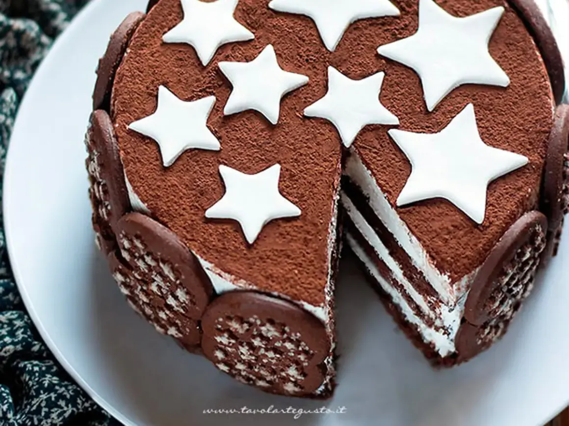 Torta Pan di Stelle ricoperta di cacao, decorata con stelle bianche e biscotti al cioccolato. Una fetta mostra gli strati soffici.