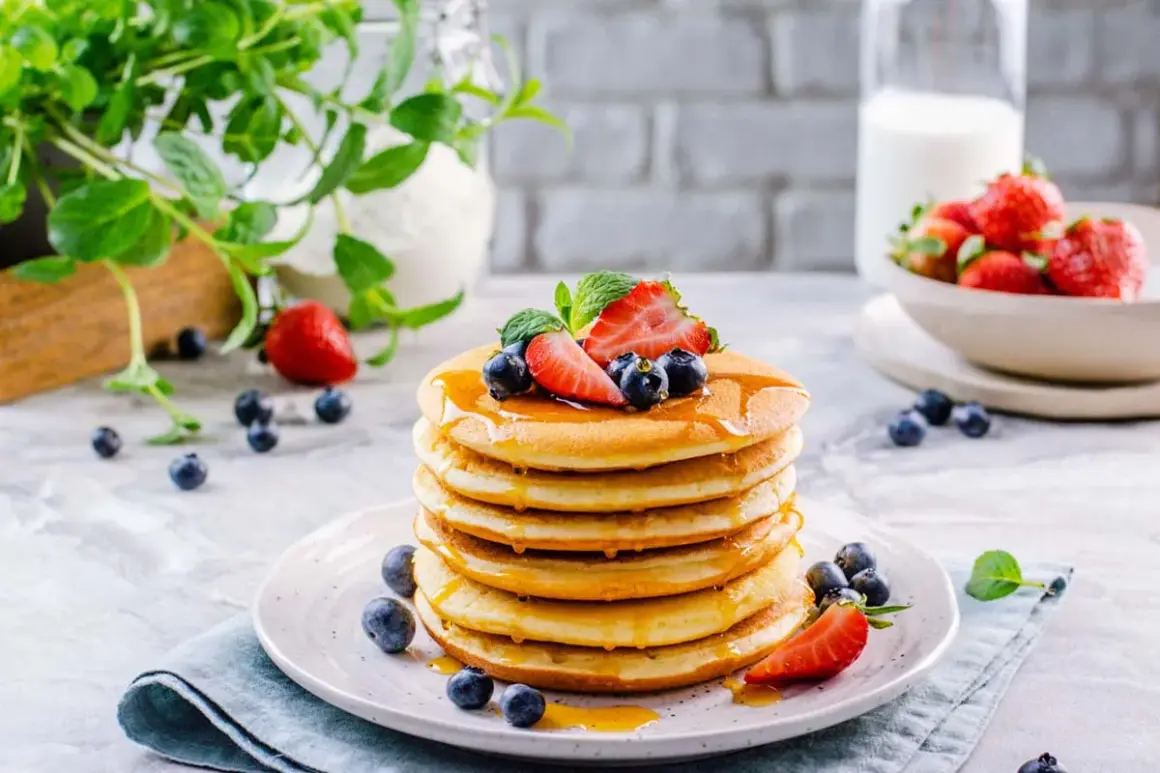 Pila di soffici pancake senza glutine e lattosio, guarniti con fragole, mirtilli e sciroppo d'acero.
