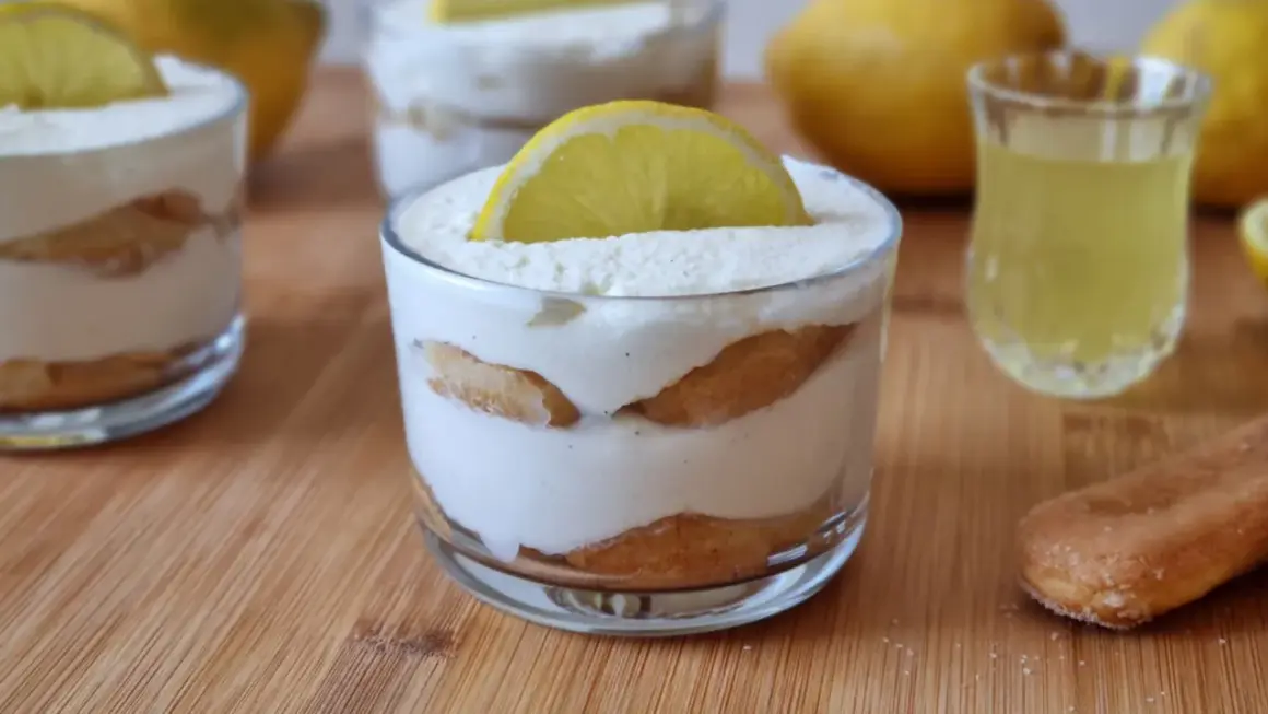 Delizioso tiramisù al limoncello in bicchieri, guarnito con una fetta di limone. Accanto, un bicchierino di limoncello e savoiardi.