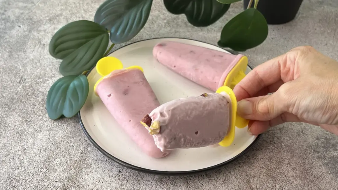 Tre ghiaccioli rosa, un gelato allo yogurt fatto in casa, su un piatto bianco con foglie verdi sullo sfondo.