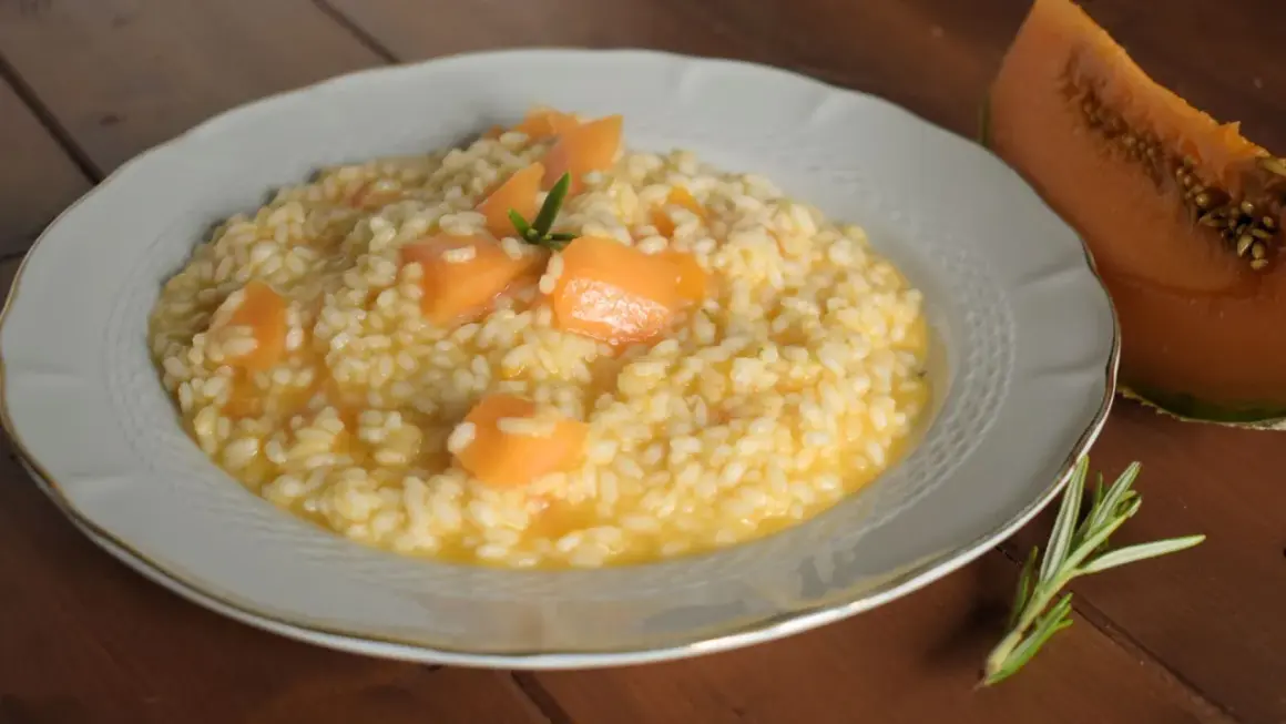 Un cremoso risotto con pezzi di melone e un rametto di rosmarino, accanto a una fetta di melone.