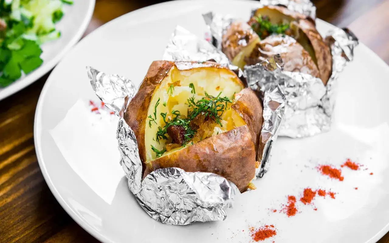 Patate al cartoccio fumanti, condite con erbe fresche e cipolla caramellata, servite su un piatto bianco con una spolverata di paprika.