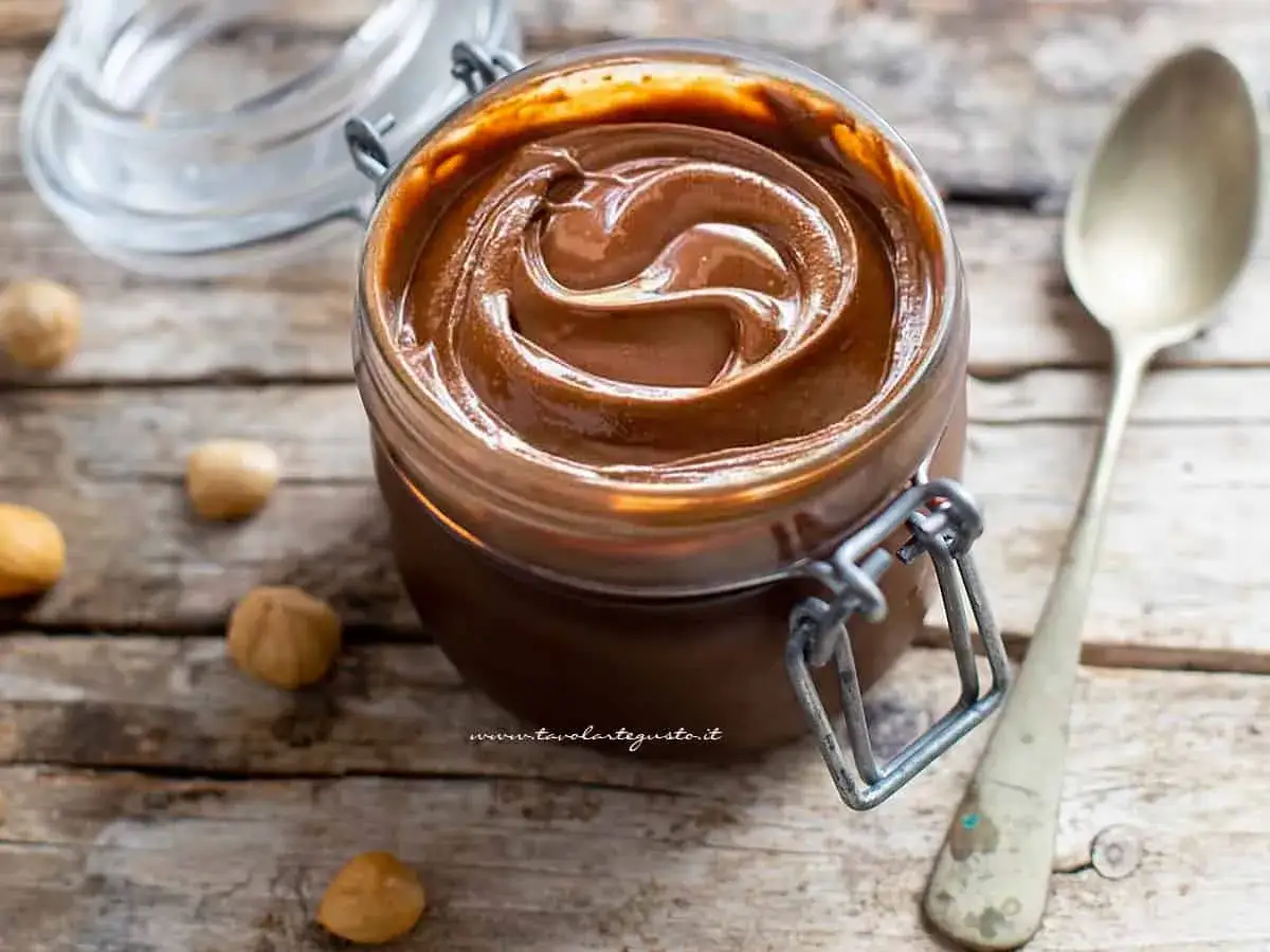 Vasetto di crema alla Nutella fatto in casa, con nocciole sparse sul tavolo di legno.