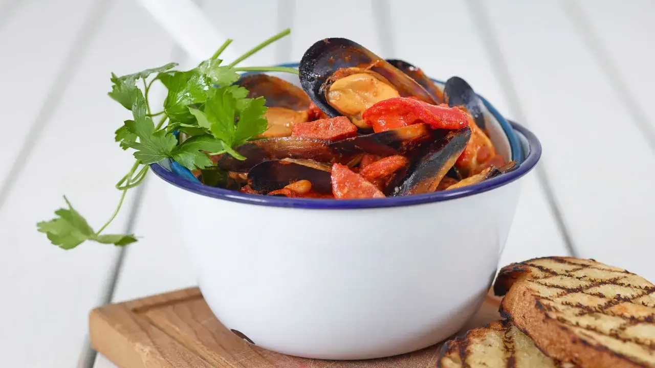 Una ciotola di zuppa di cozze al pomodoro, guarnita con prezzemolo fresco e accompagnata da pane grigliato.