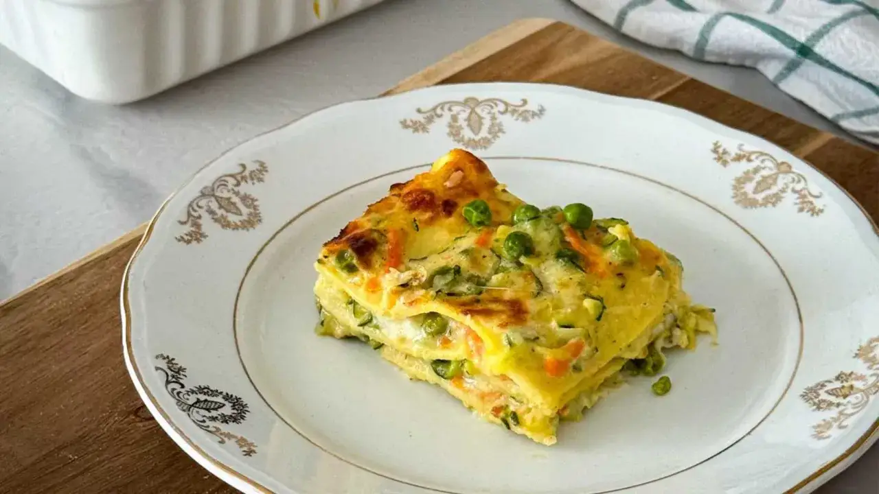 Una fetta di lasagne vegetariane gustose, con piselli e zucchine, servita su un piatto bianco con decorazioni dorate.