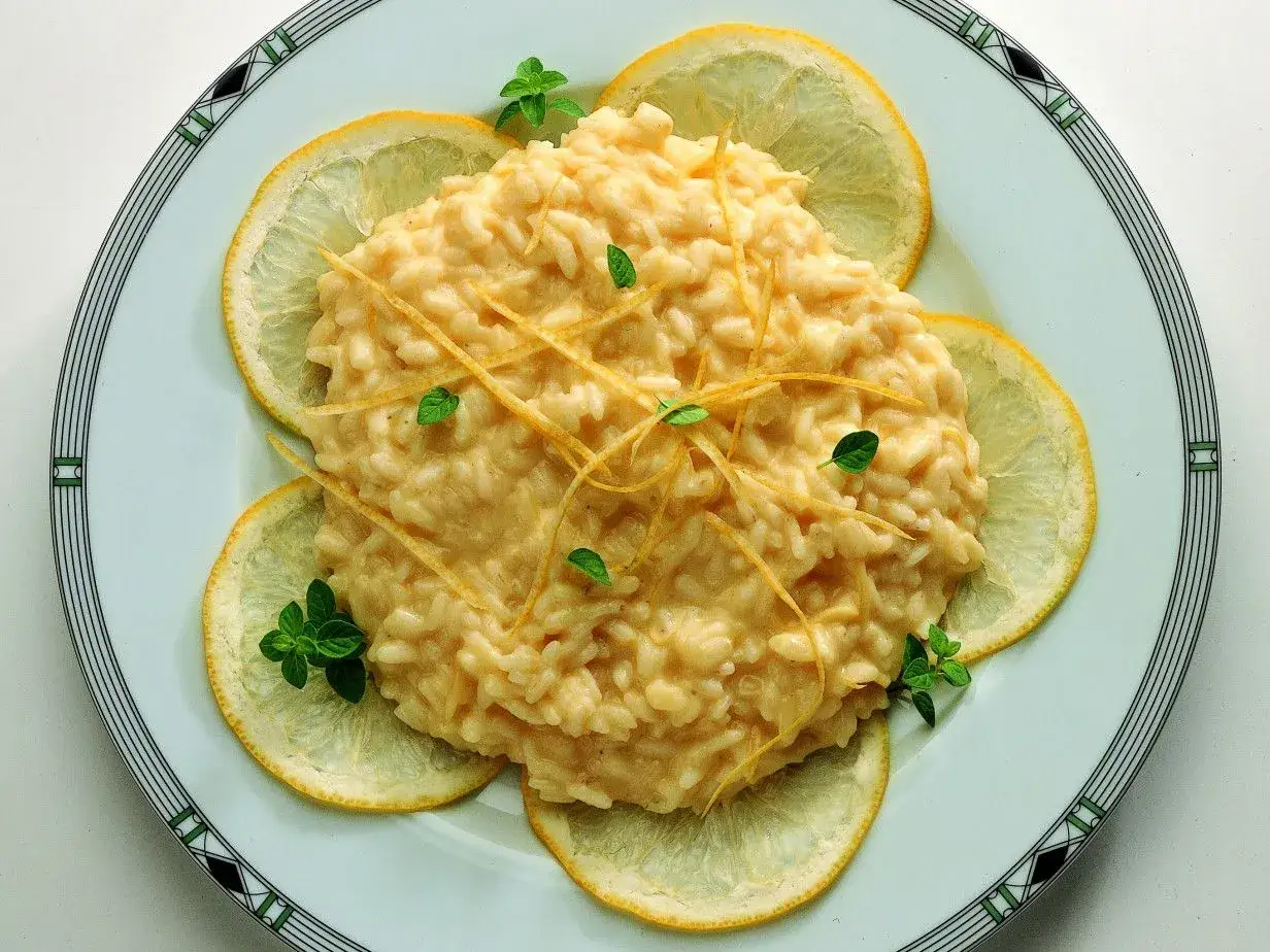 Un cremoso risotto al limone, guarnito con scorzette e foglioline verdi, circondato da fette di limone fresco su un piatto elegante.