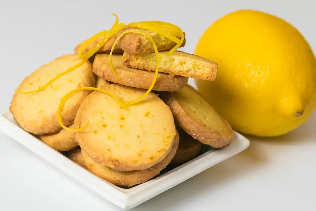 Biscotti senza burro al limone, dorati e profumati, guarniti con scorzette fresche. Accanto, un limone intero, pronto a donare il suo aroma.