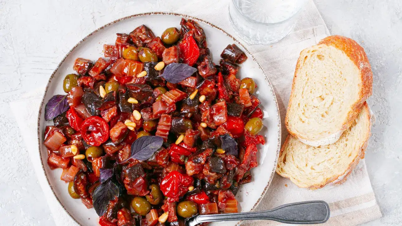 Un piatto invitante di caponata di verdure, con melanzane, pomodori, olive e pinoli, accompagnato da fette di pane fresco.