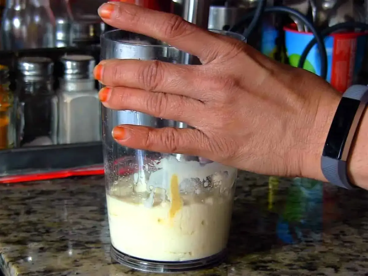 Mano che prepara maionese senza uova con un frullatore a immersione. La crema è già quasi pronta.