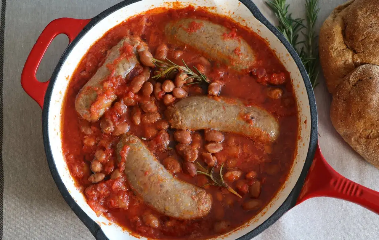 Salsiccia e fagioli in umido, un piatto rustico e saporito, servito in una padella rossa con rosmarino e pane integrale.