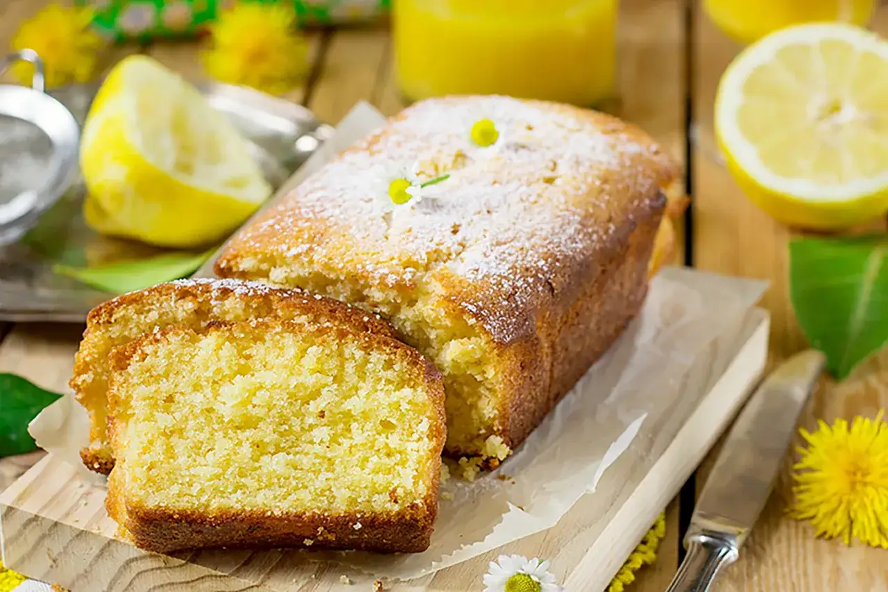 Un soffice plumcake al limone, spolverato di zucchero a velo, con fette di limone e fiori gialli sullo sfondo.