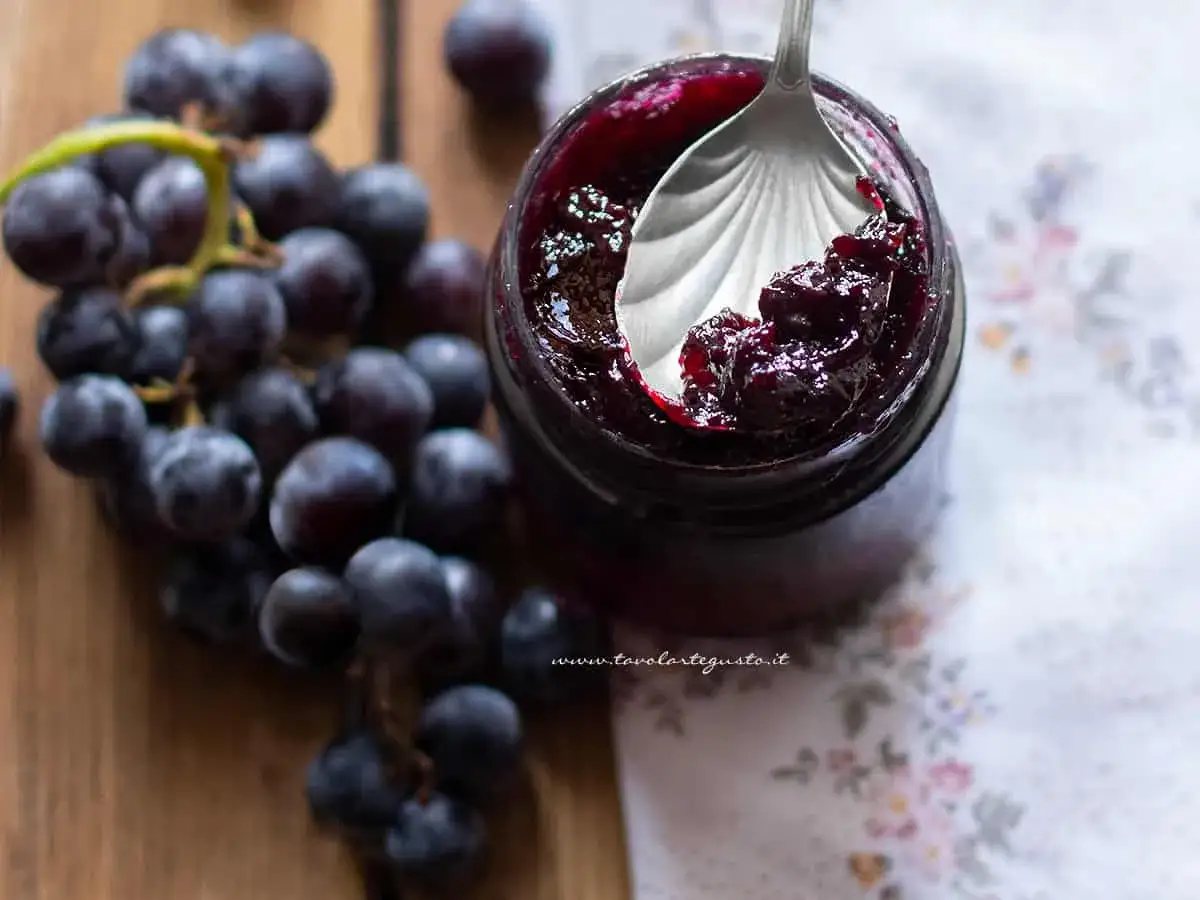 Marmellata di uva con buccia, scura e invitante, in un vasetto, con un grappolo d'uva nera accanto.