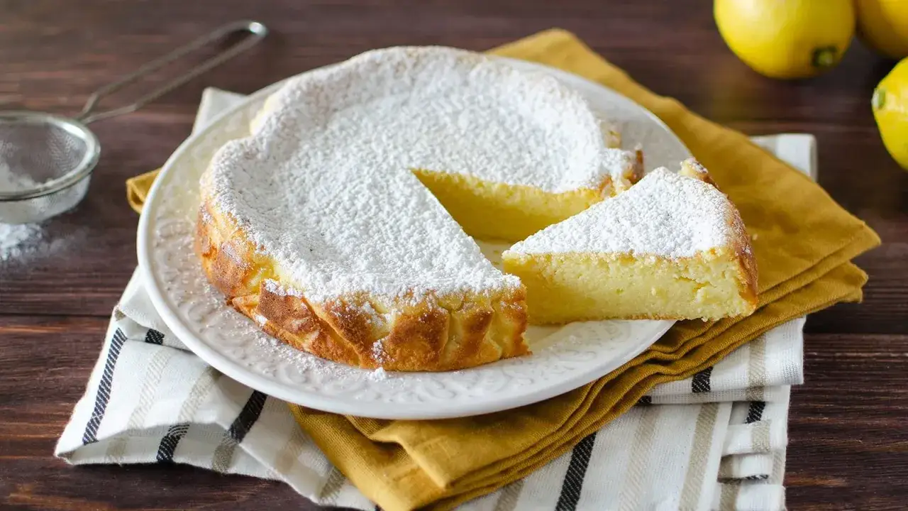 Deliziosa torta di ricotta spolverata di zucchero a velo, con una fetta tagliata. Accanto, limoni freschi e un setaccio.