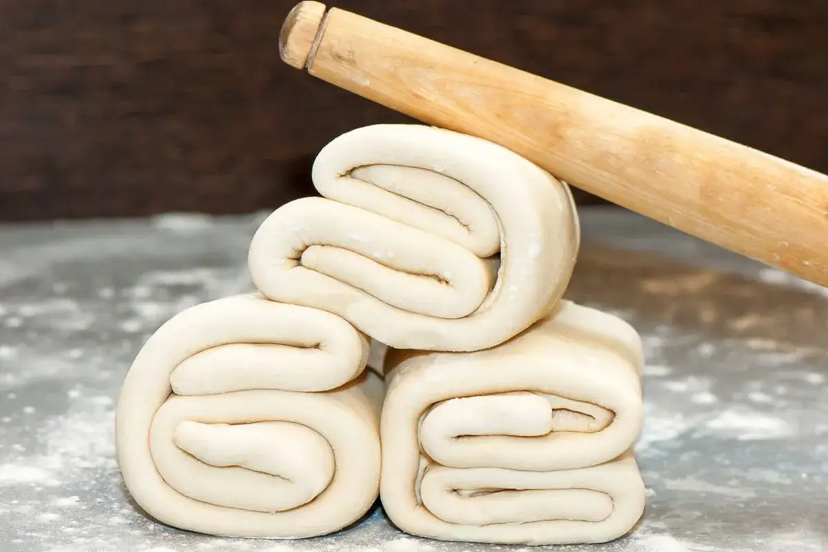 Impasti arrotolati per una ricetta pasta sfoglia veloce senza riposo. Un mattarello in legno &egrave; appoggiato sopra.