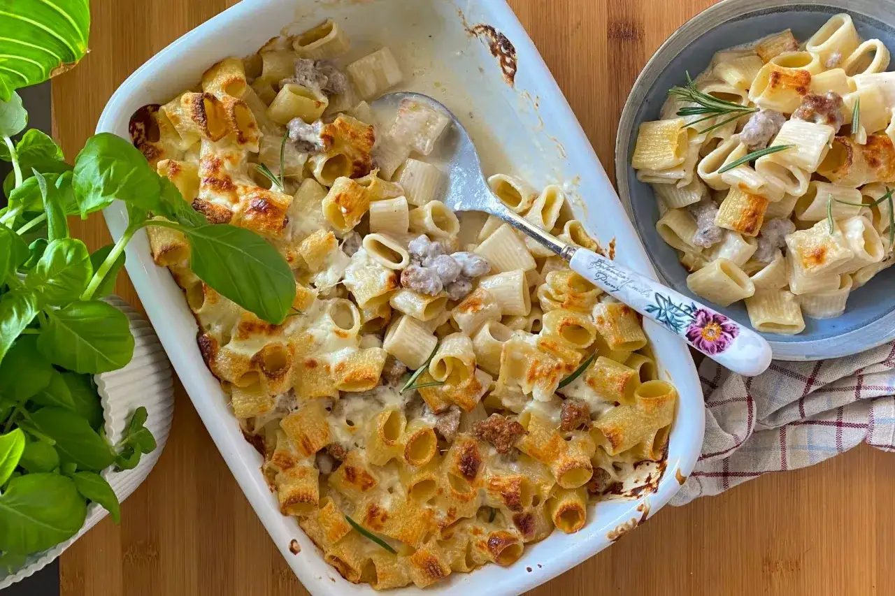 Delizioso pasta al forno con salsiccia, dorata e filante, servita in una teglia bianca e un piatto blu, con basilico fresco a lato.
