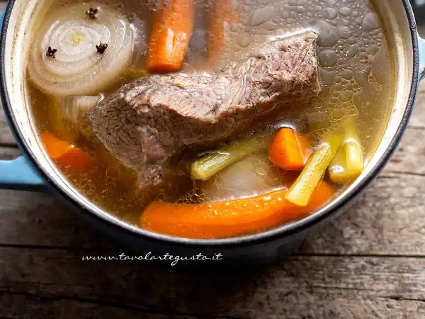 Brodo di carne ricetta: un pezzo di carne, carote, sedano e cipolla con chiodi di garofano bollono in una pentola.