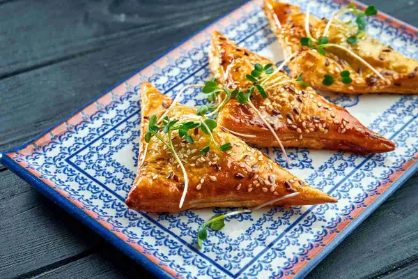 Triangoli dorati di pasta fillo ricetta, cosparsi di semi di sesamo e guarniti con germogli freschi, su un piatto decorato.