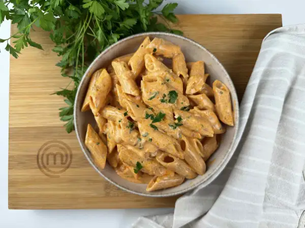 Penne alla vodka cremose e invitanti, guarnite con prezzemolo fresco, su un tagliere di legno con il logo MasterChef.
