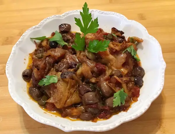 Un piatto invitante di melanzane in agrodolce, con olive nere e capperi, guarnito con prezzemolo fresco.