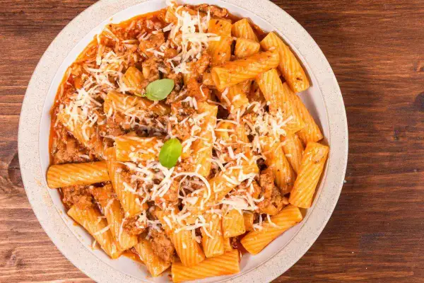 Un piatto di pasta alla zozzona fumante, con rigatoni conditi da un ricco ragù e abbondante parmigiano grattugiato, guarnito con basilico fresco.