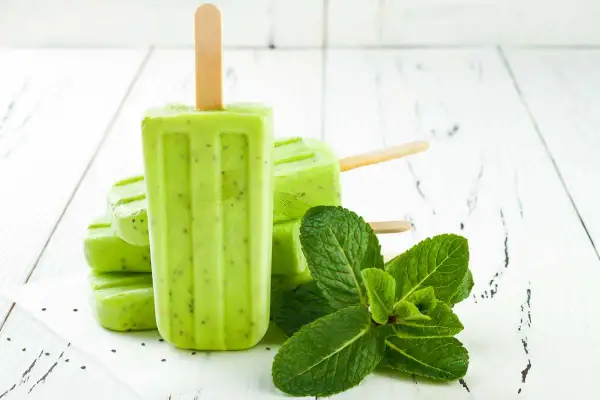 Ghiaccioli alla menta, freschi e verdi, con semi di kiwi visibili, accanto a foglie di menta fresca su un tavolo di legno bianco.